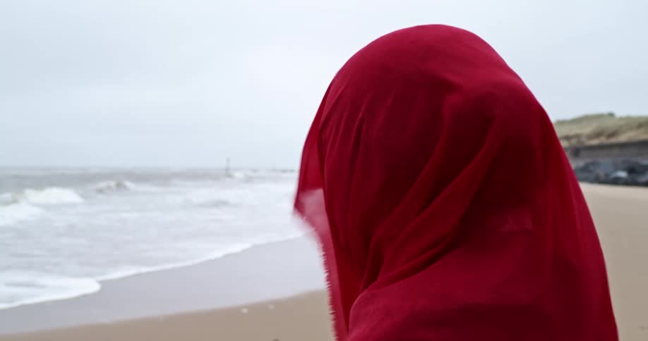 A woman in a head scarf stands on a beach on a windy day