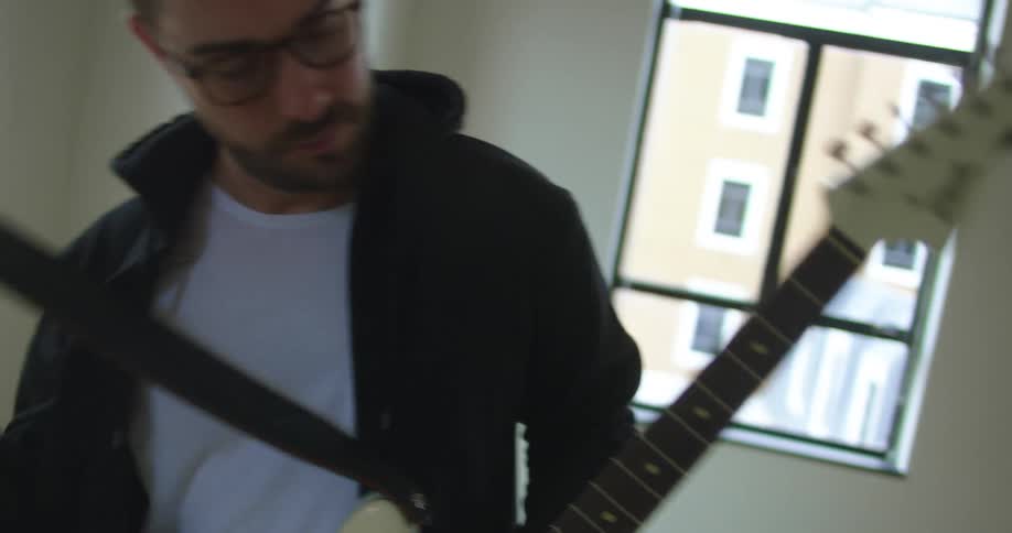 Man with stubble beard and glasses picks up a guitar to play in a room