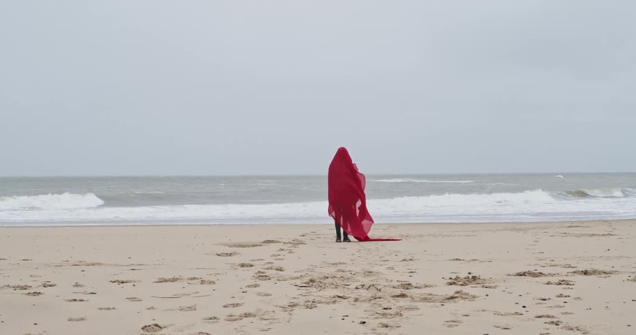 Woman wearing red fabric stands on the beach as she looks out at the ocean