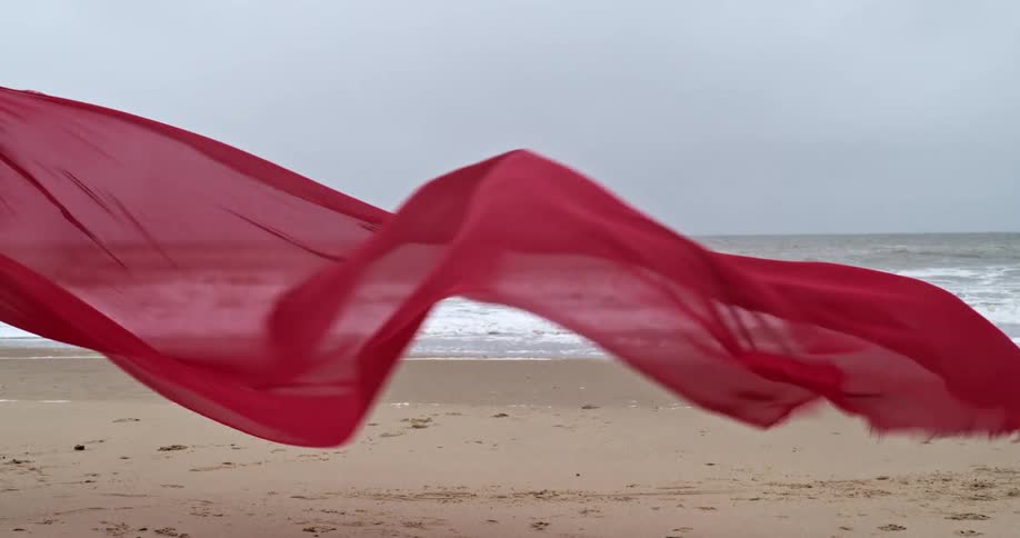A red see through cloth blowing in the wind on the beach with the ocean and the sky in the background