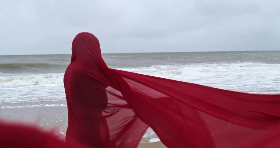 A woman wearing only a sheer red fabric that billows in the wind walks along a beach just out of reach of the waves on a cloudy day