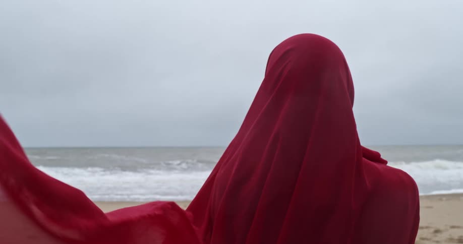 A person with a head covering standing on a beach with the sky and the ocean in the background