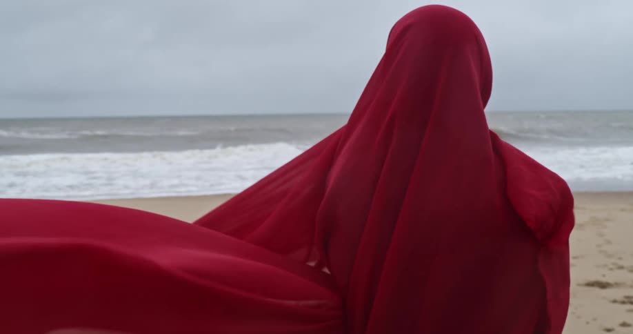 A person covered in a red cloth walking on the beach with the ocean sky and clouds in the background