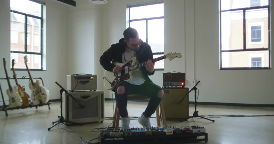 A man sits in a chair playing an electric guitar while surrounded by musical equipment