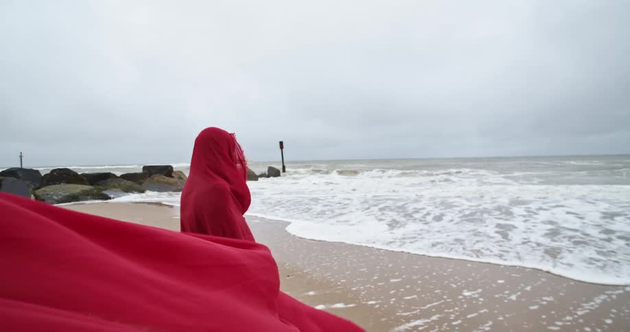 A woman completely covered up in a cloak walks towards the water