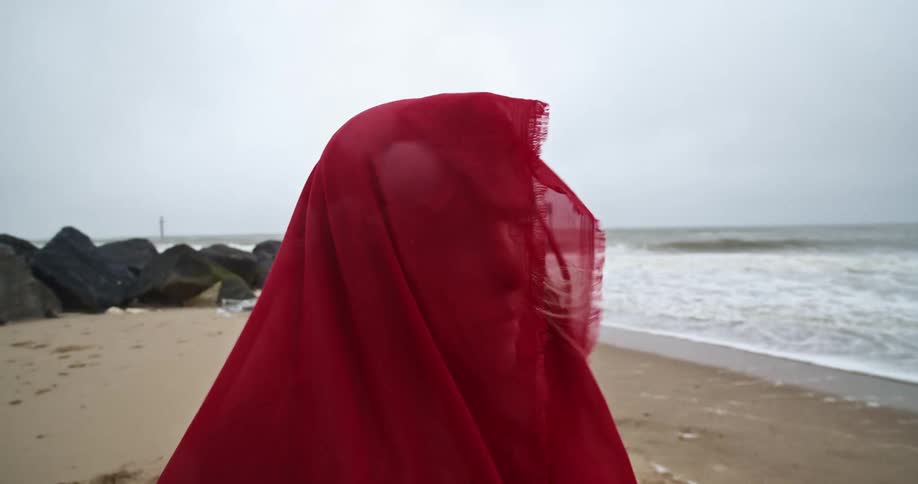 Blonde woman wearing a red scarf stands on the beach next to the sea on a cloudy day