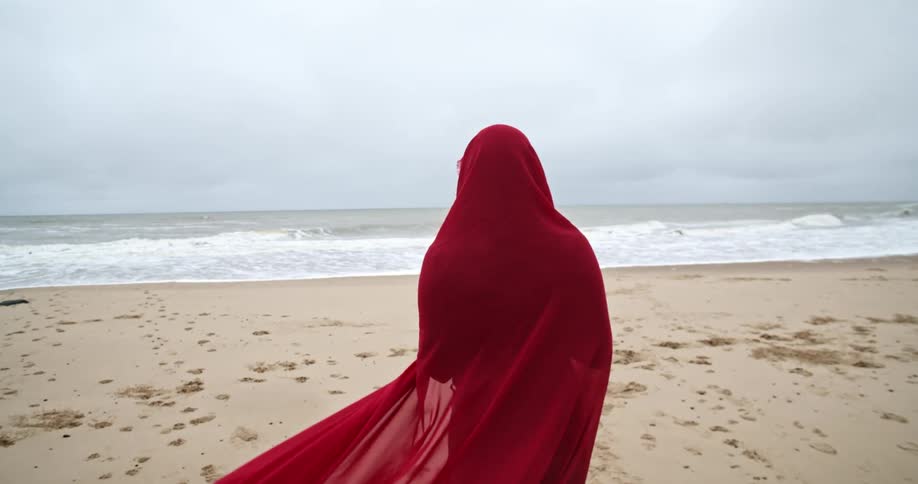 A woman wrapped in a billowing sheer red scarf walks toward the ocean waves