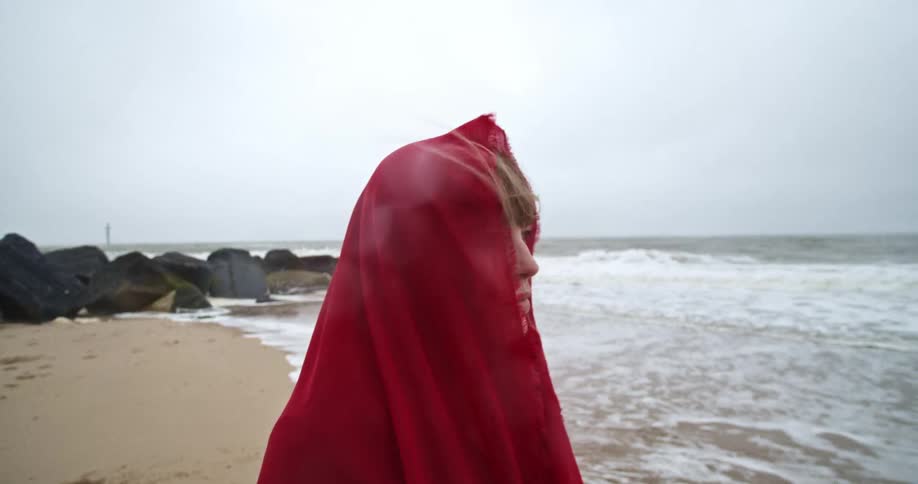 Blonde woman wearing a red veil stands on the beach next to the ocean on a cloudy day