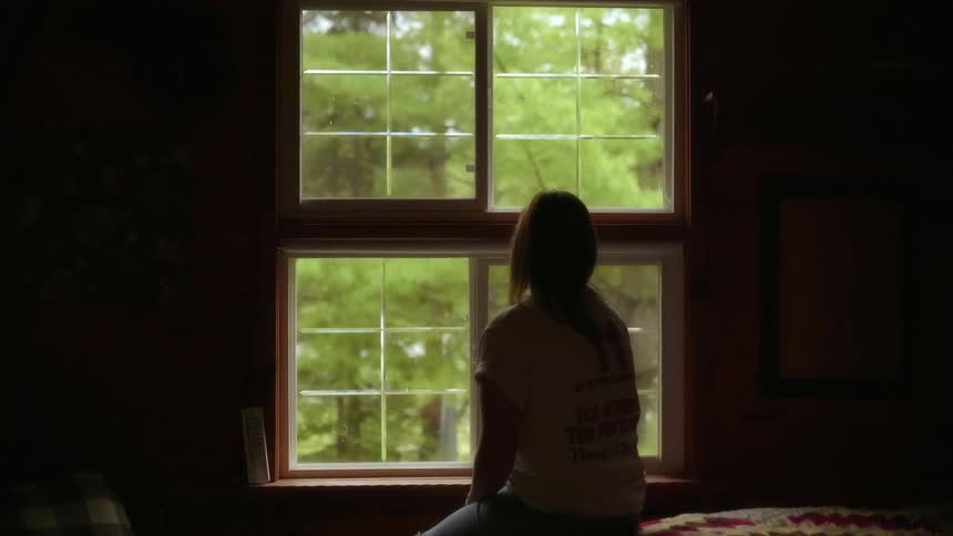 Woman sitting on a bed looks out the window at green trees in a dark room