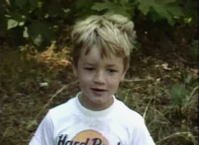 A boy with a t shirt and a fanny pack standing and talking outside with plants in the background