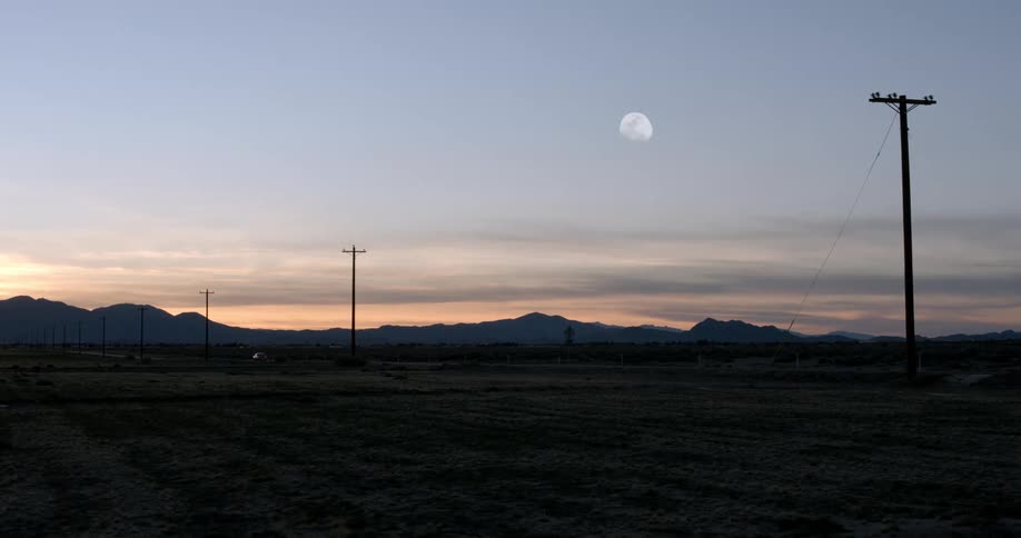 Car drives down a road through the desert under the moon at sunset