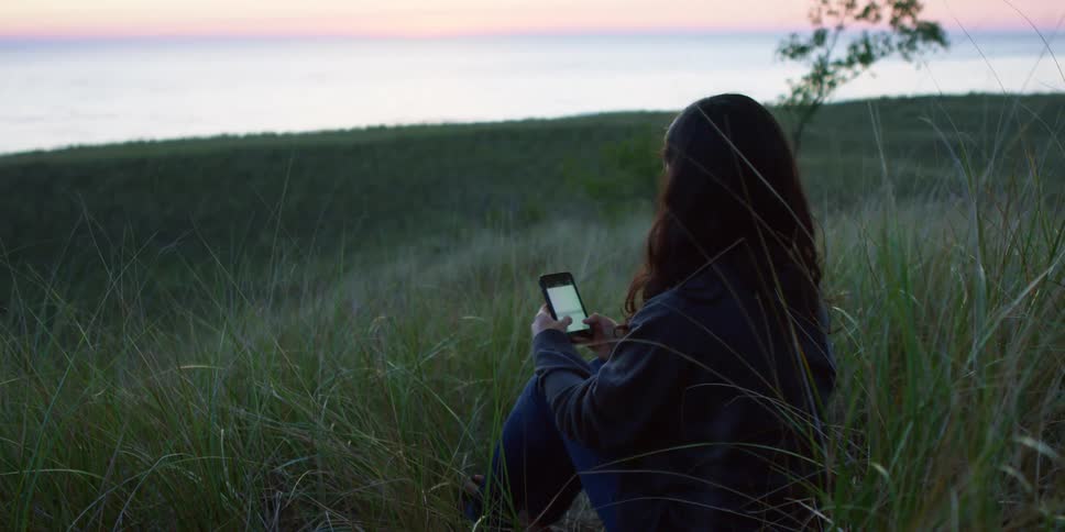 A woman types on her phone while sitting in a field with tall grass
