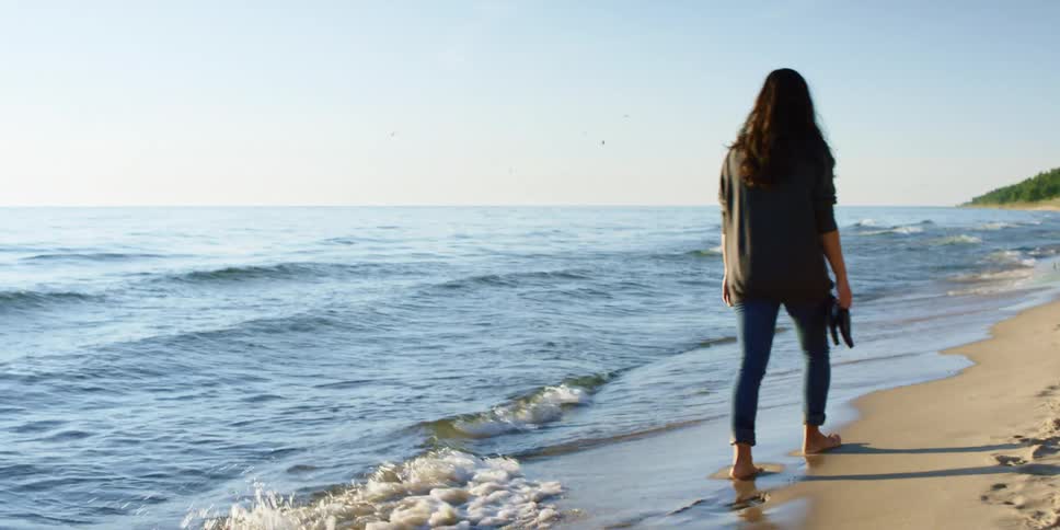 A woman walks barefoot by the waves carrying her flip flops on a bright sunny day