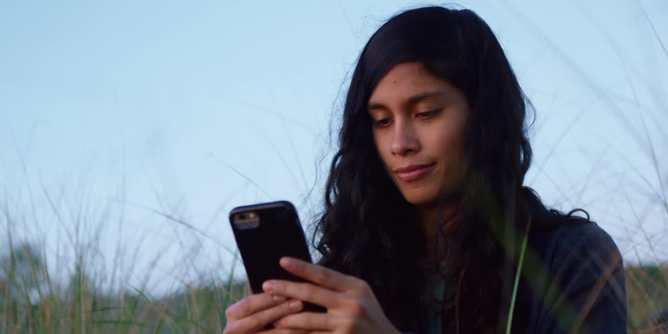 Brunette teenage girl with black curly hair sitting on the grass uses a smart phone at dusk