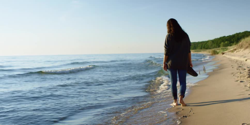 Woman walks along beach near water carrying shoes