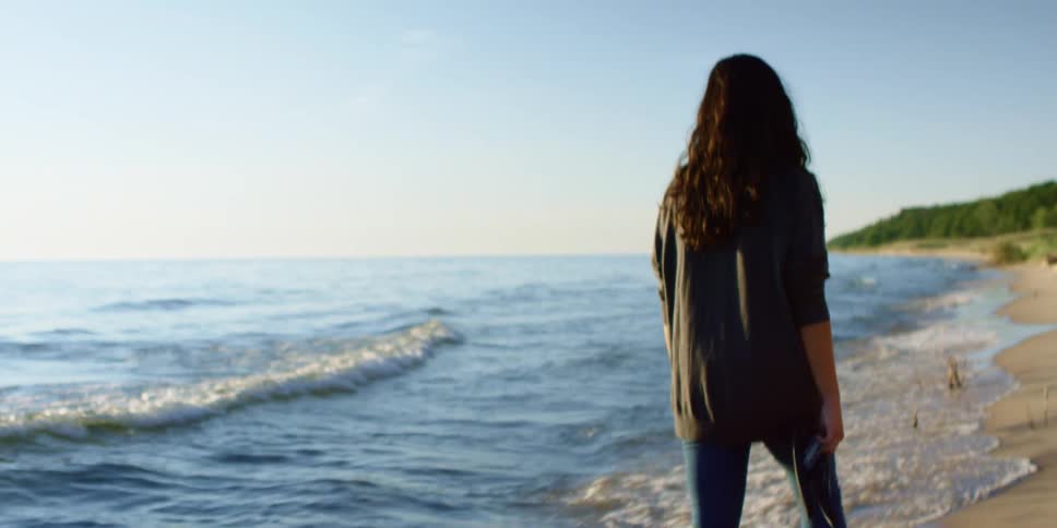A woman carries her sandals as she slowly walks along a beach just out of reach of the ocean waves