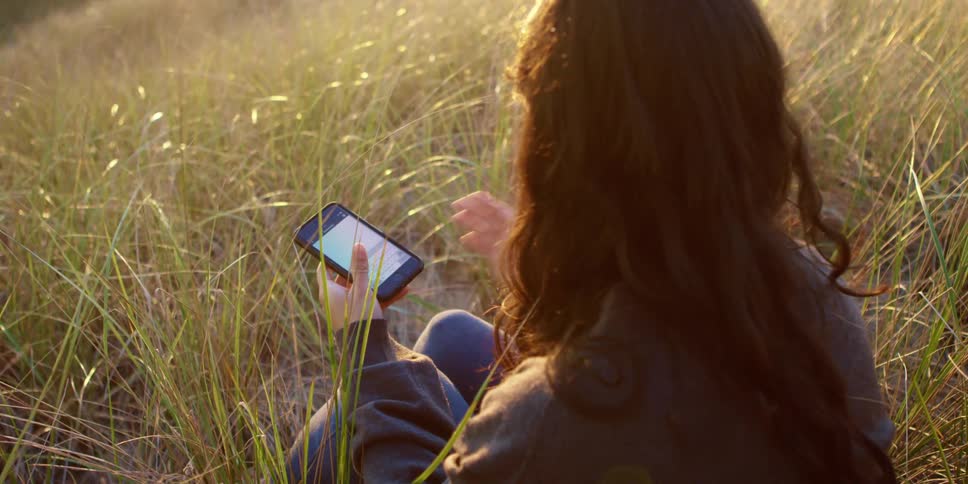 A woman is sitting in a grassland area texting on a cell phone