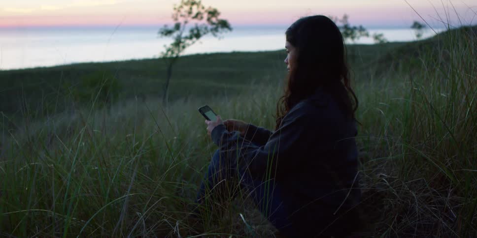 Woman sits on ground in field near shore texting with red sky near the horizon