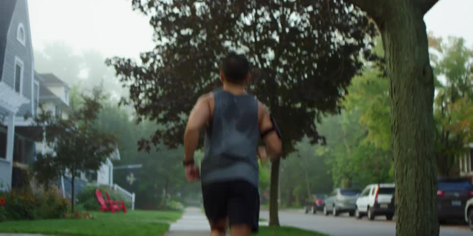 Male standing on sidewalk in a suburban neighborhood with an electronic device strapped to his arm begins to jog down the street