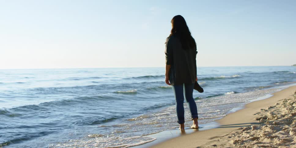 Brunette girl with curly black hair stands barefoot on the beach and watches the sea