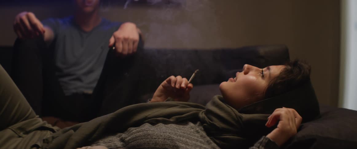 Woman with a head scarf lies down on a sofa and smokes a cigarette in front of a man in the living room at home