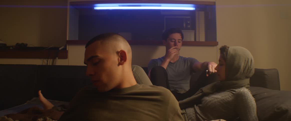 Three teenagers relax on a couch inside a house as one boy lights a cigarette and starts to smoke