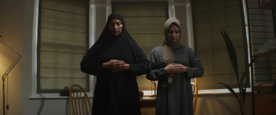 Two women with head coverings touching their heads with a table chairs and windows in the background