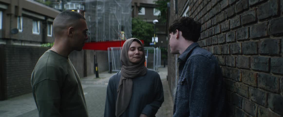 A woman in an alleyway kissing a man and giving another man a hug with trees and buildings in the background