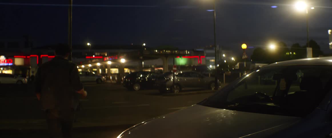A teenager leaning against his car in a parking lot looks at his phone and then walks away