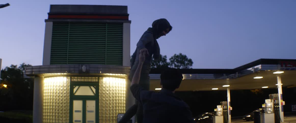 A woman walks atop a traffic barrier while holding the hand of a man walking on the ground beside her before the man helps her to the ground