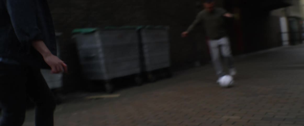 Two men and a woman with brown head scarf play football in the narrow street