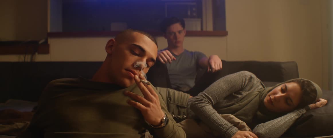 Two men and a woman in a room the woman hands the man a cigarette and he smokes it