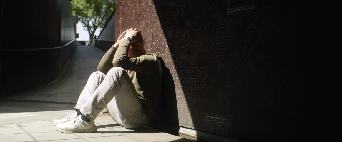 A teenager sitting on a sidewalk and leaning against a building puts his head in to his hands