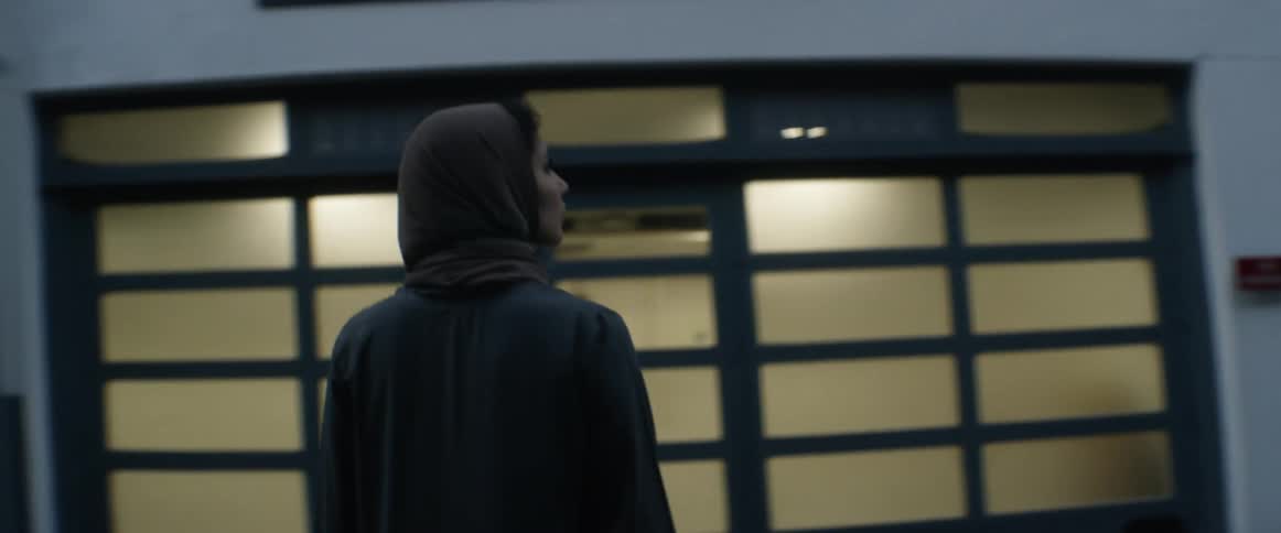 A woman stands on a street between tall buildings looking up at the buildings around her