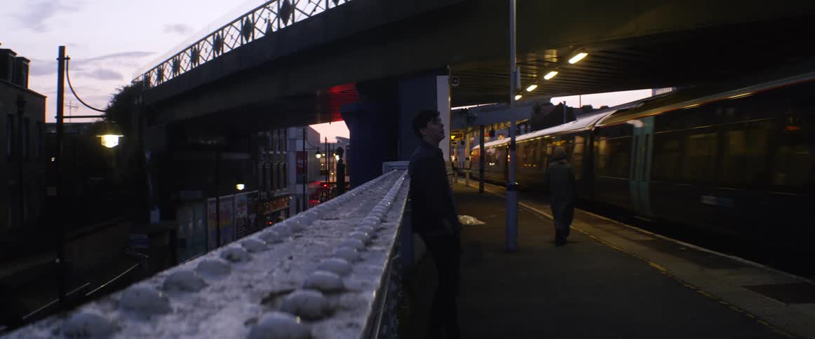 People waiting in a train station as a train arrives with the sky and clouds in the background