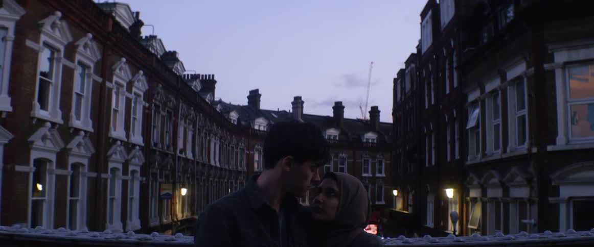 A young man and woman cuddle and kiss outside an apartment building