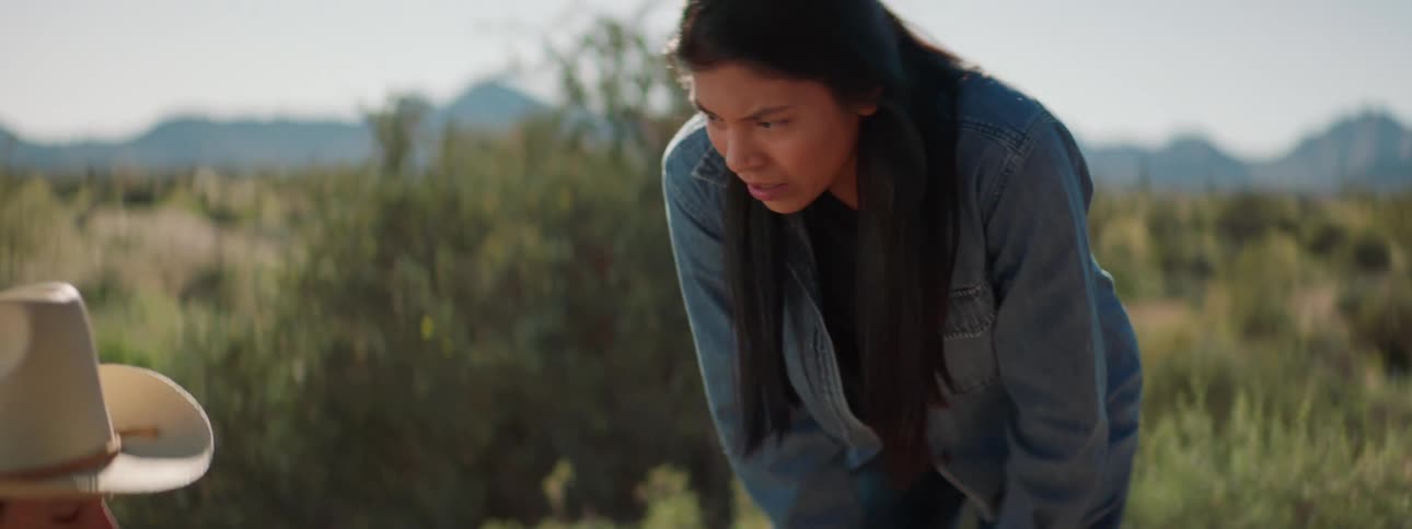 A woman bends down to a man sitting on the ground in a very rural setting