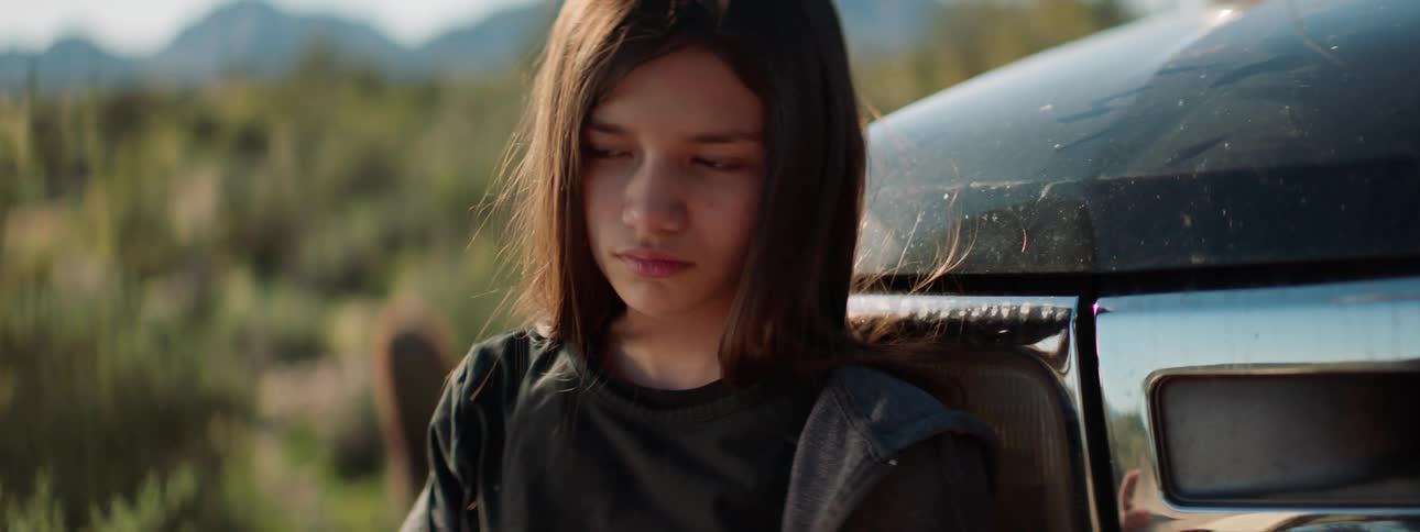 A girl standing in front of a vehicle reading a book with the desert and mountains in the background