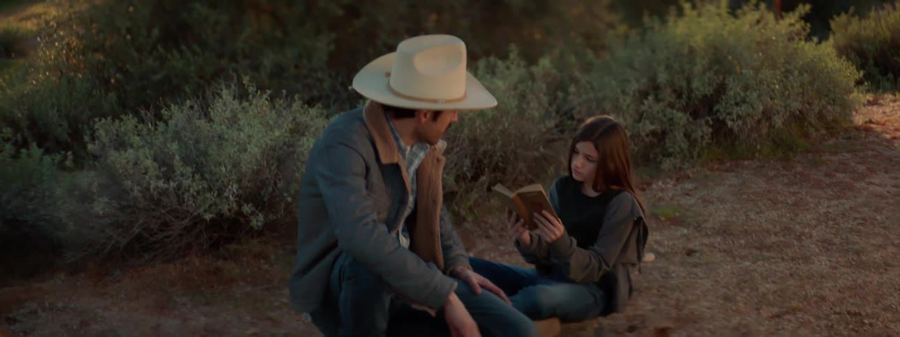 Man wearing cowboy hat sits next to girl reading book in the dirt at the side of the road