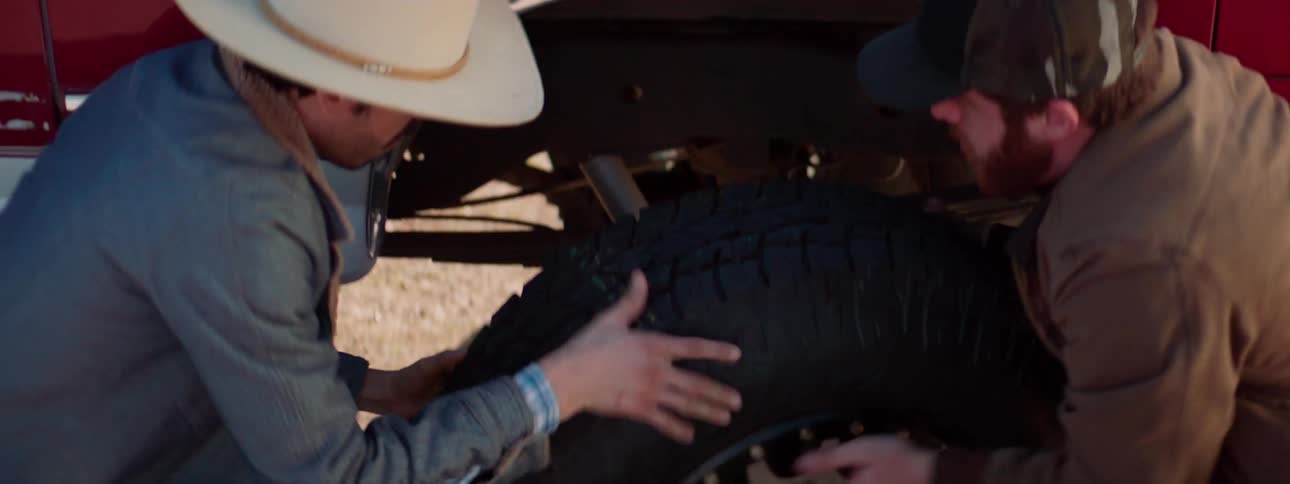 Two men remove a tire from a red and white truck on a dirt road