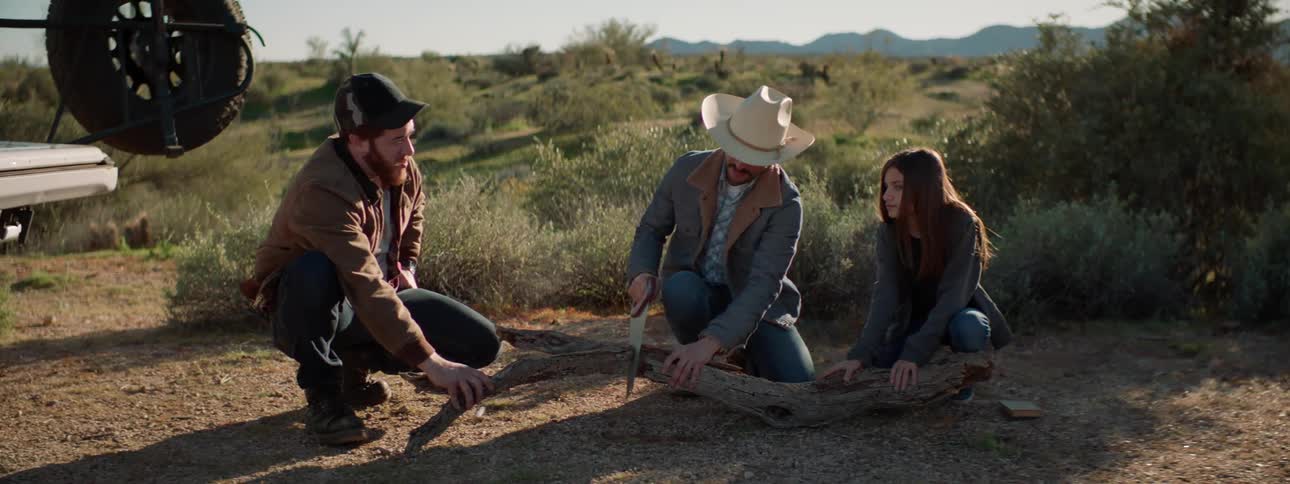 A man with a saw and a cowboy hat cutting a log next to another man and woman