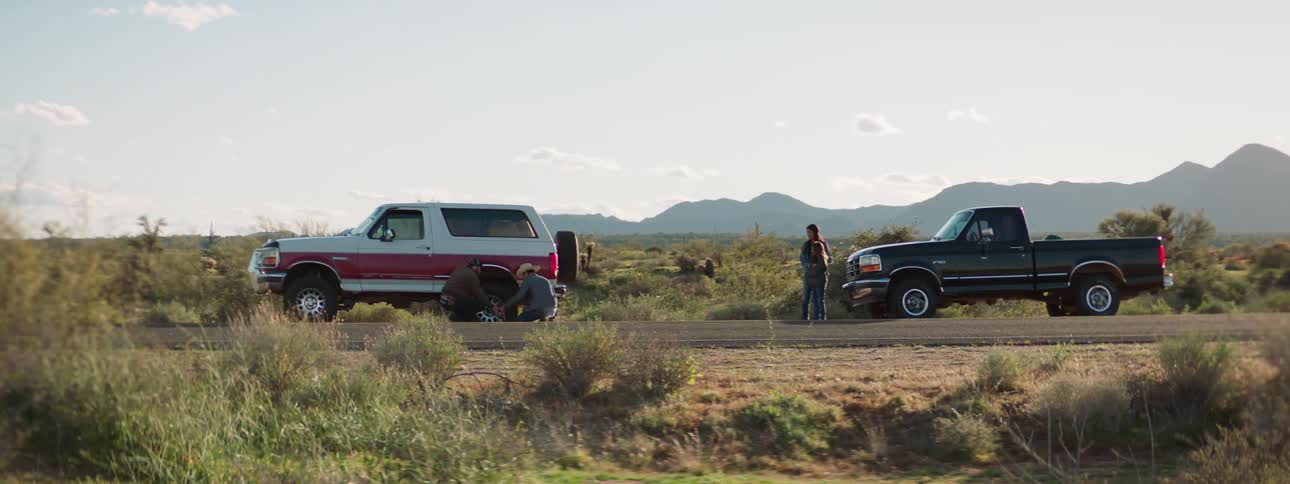 Men change tire on shoulder of highway near scrub land andmountain while girl and woman wait