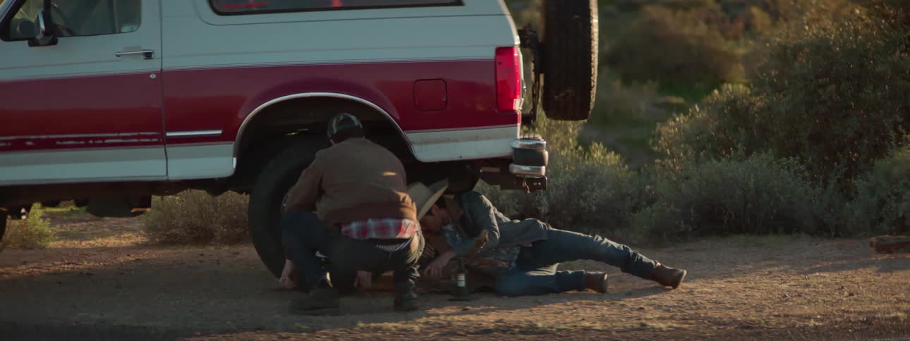 Two vehicles are stopped at the side of a road and a man walks away from two girls to go help another man change a tire