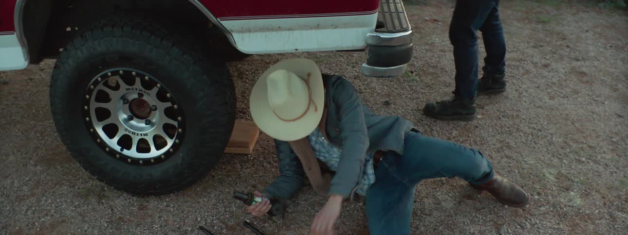 A man changes a tire on a vehicle and then walks to a truck where a teenager and young girl wait