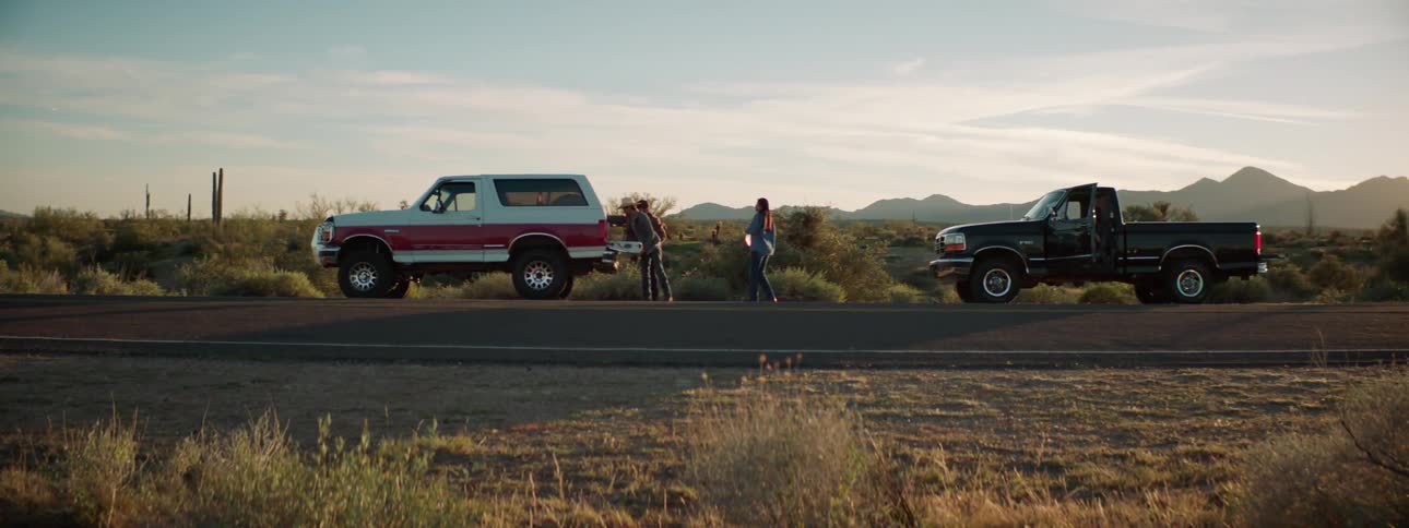 Two men putting a spare tire in the back of a truck while a woman takes a bottle of water from another truck in the desert