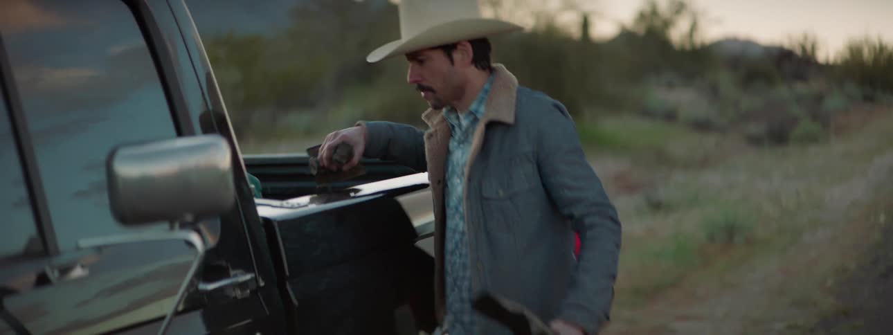 A man wearing a cowboy hat takes a rifle from the back of his truck in a rural setting