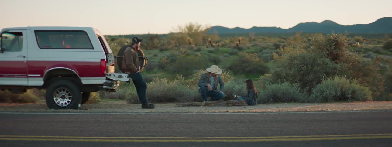 Men sit talking to woman sitting on side of road near scrub land with mountains on the horizon