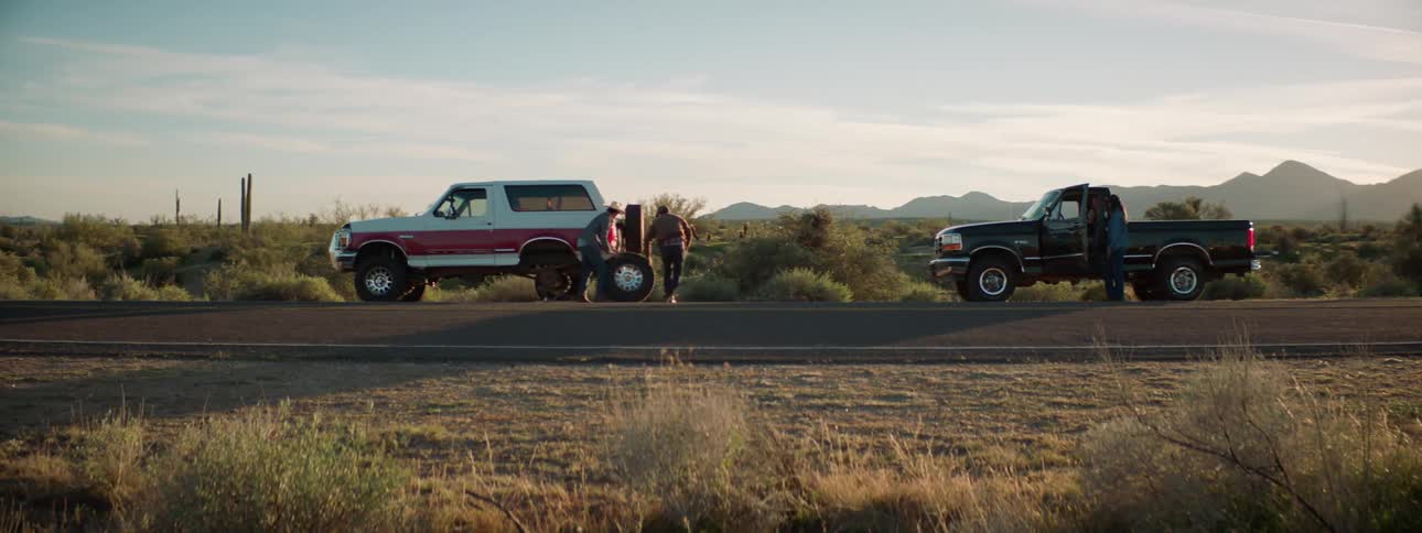 Two people change a tire on a vehicle as a teenager stands by another truck