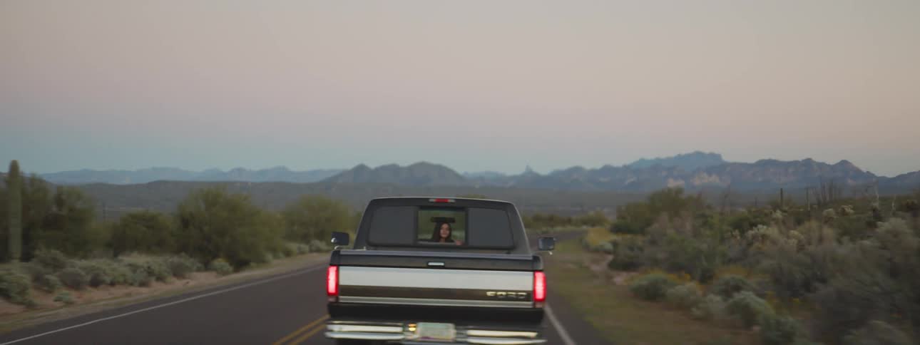 A girl looks out the rear window of a truck traveling through the countryside into the camera following behind