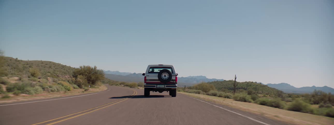 A vehicle drives down an empty two lane highway surrounded by cacti and shrubs